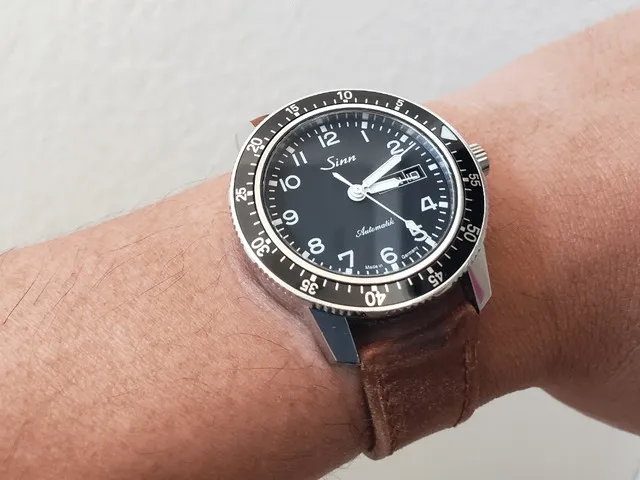 Sinn 104 black dial diver on brown leather strap, wrist shot against white wall.