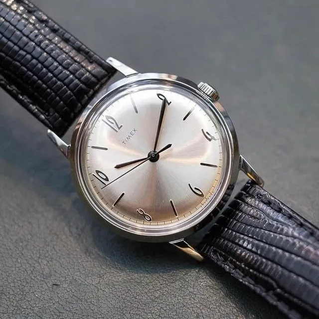 Timex vintage-style silver sunburst dial on black leather strap, slight three-quarter angle.