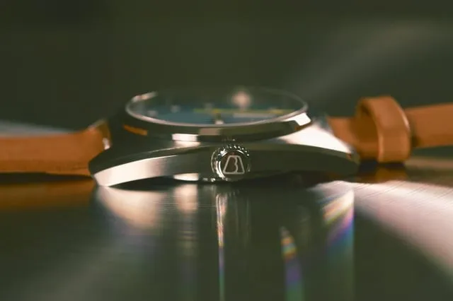 Side profile of steel-cased watch showing prominent signed crown against case flank, tan strap