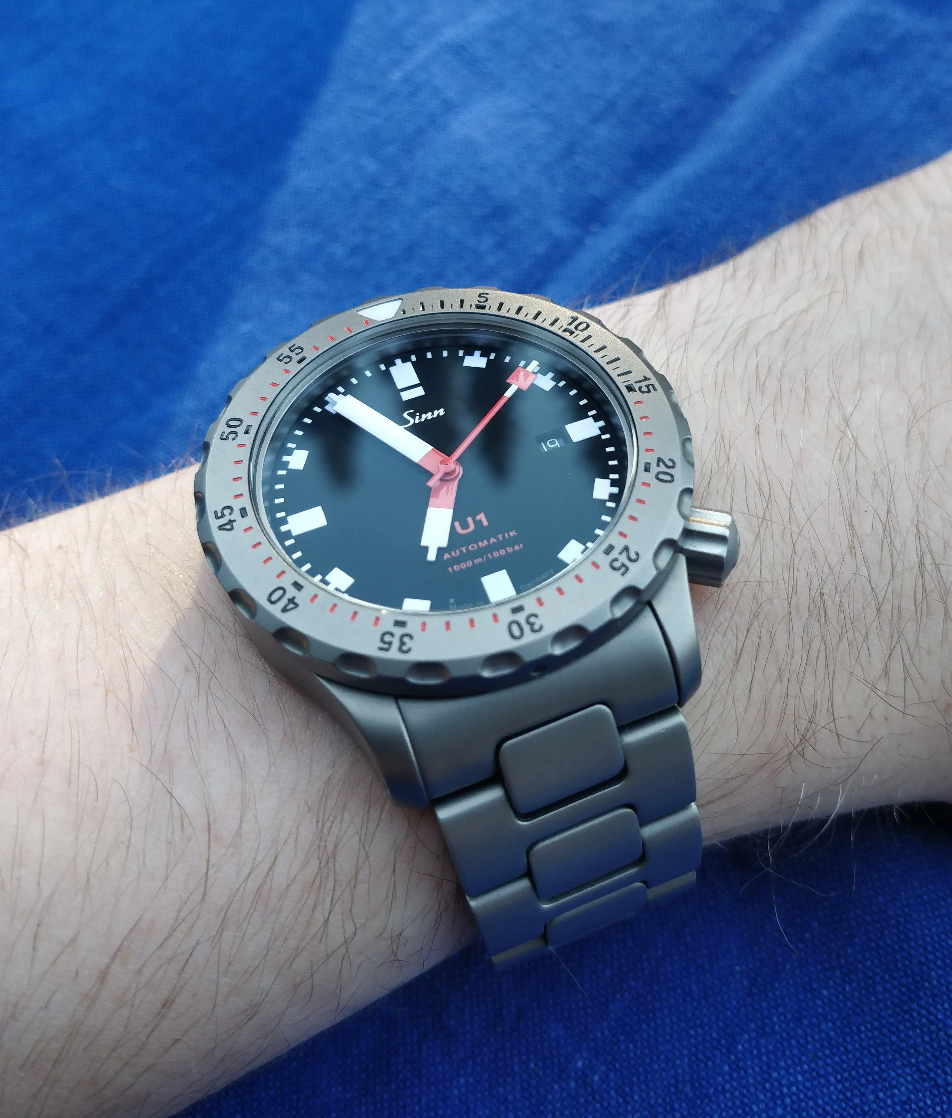Sinn U1 on wrist, black dial, red hands, tegimented steel bracelet, blue background.